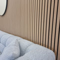 Close-up of a wood acoustic slatted wall panel with vertical slats, installed behind a grey upholstered sofa in a room with a light wooden finish.