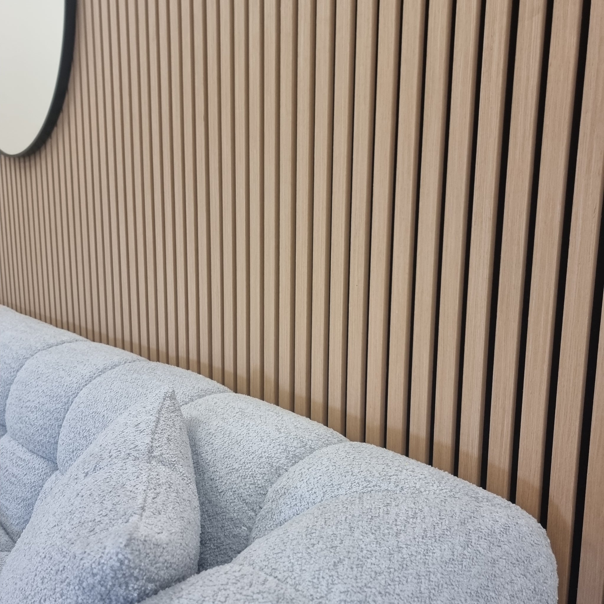 Close-up of a wood acoustic slatted wall panel with vertical slats, installed behind a grey upholstered sofa in a room with a light wooden finish.